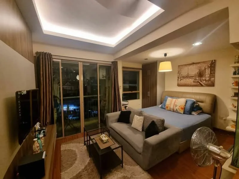 Living features: grey fabric sofa, coffee table, wall-mounted TV, wooden TV console, area rug with geometric pattern, floor-to-ceiling windows, curtains, ceiling with recessed lighting, decorative wall art, wood panel accent wall, fan, queen-size bed with upholstered headboard; style: modern; view: pool.