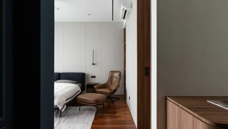 Bedroom features: king-size bed with white bedding, upholstered headboard, leather armchair, ottoman, wooden flooring, wall-mounted reading light, air conditioning unit, decorative wall paneling, neutral wall color, area rug; style: modern. Room features: wooden cabinetry, unknown countertop.