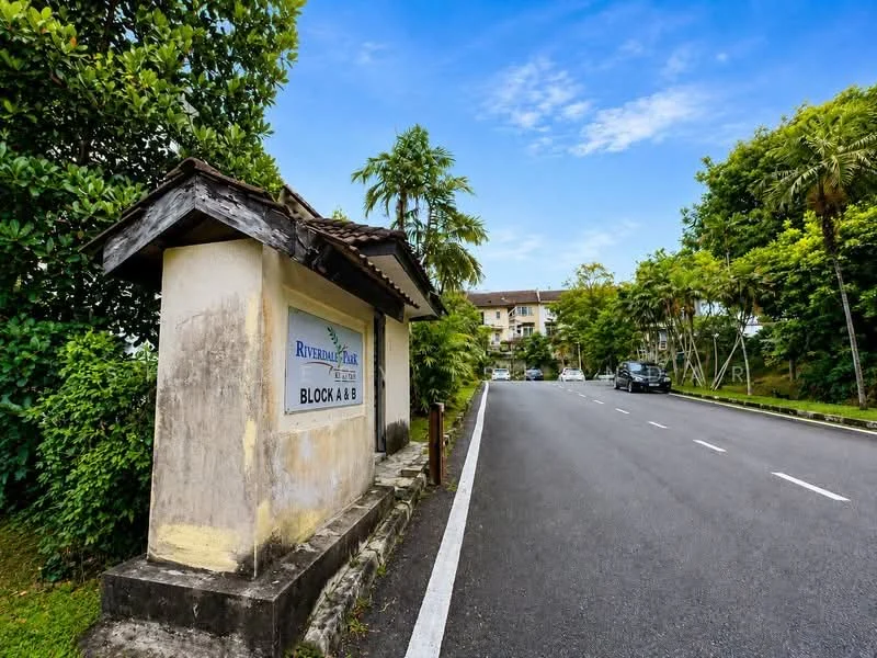 Exterior type: low-rise apartment block; features: signage with 'Riverdale Park', tree-lined street, paved road, landscaped greenery, parking spaces visible, two-storey building in background, tropical landscaping, clear blue sky; style: modern.