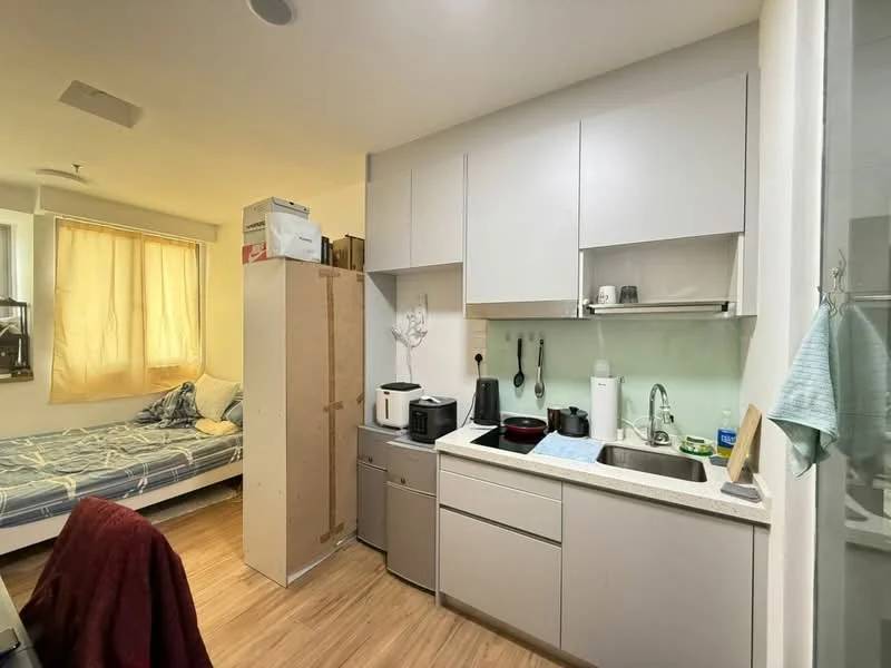 Kitchen features: light grey cabinetry, white countertop, single-bowl stainless-steel sink, chrome pull-down faucet, built-in stove, microwave, refrigerator, wooden flooring, window with sheer curtains, kitchen island, appliances on countertop; style: modern. Room features: bed with blue bedding, wall-mounted shelf, light-colored walls, natural light from window.