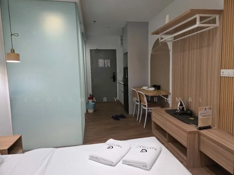 Room features: wooden flooring, light wood paneling, white walls, glass partition, desk with chair, wall-mounted shelf, bed with white linens, hanging pendant light, door to hallway, small kitchenette area, towels on bed, wall-mounted mirror; style: modern.
