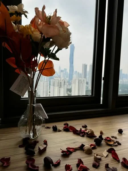 Room features: vase with flowers, wooden table surface, decorative petals on table, large window, city skyline view, glass vase; style: modern; view: city.