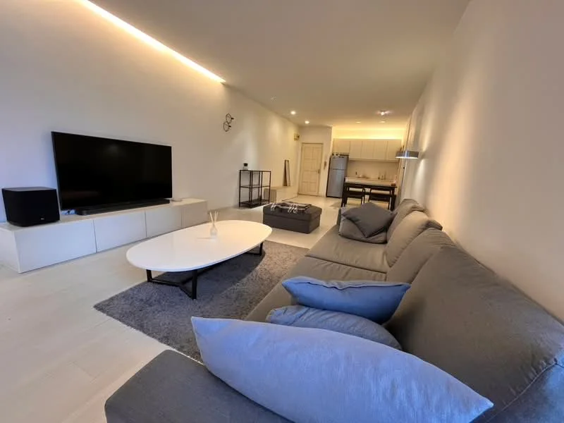 Living features: 3-seater fabric sofa in grey, coffee table, TV console, wall-mounted TV, area rug, recessed LED downlights, light-colored walls, light-colored flooring, decorative wall clock, open layout to kitchen, blue decorative cushions; style: modern. Kitchen features: kitchen island, stainless-steel fridge, built-in cabinets, countertop, open layout to living area; style: modern.