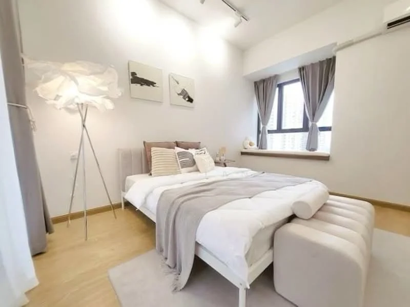 Bedroom features: queen-size bed with white bedding, upholstered headboard, bedside lamps, light grey throw blanket, decorative pillows in various colors, white area rug, floor-to-ceiling window with sheer curtains, wooden flooring, wall-mounted air conditioning unit, framed artwork on walls, light fixture with track lighting, window seat with cushion; style: modern; view: city.