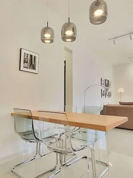 Dining features: transparent acrylic dining chairs, wooden dining table with light wood finish, three pendant lights with metallic shades, white painted walls, glossy tile flooring, framed artwork on walls, open layout to living area; style: modern.