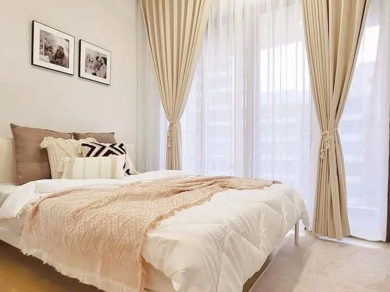 Bedroom features: queen-size bed with white bedding, decorative throw blanket in peach, two decorative pillows, framed artwork on walls, floor-to-ceiling windows with sheer curtains, beige curtains, wooden flooring, area rug with geometric pattern; style: modern.