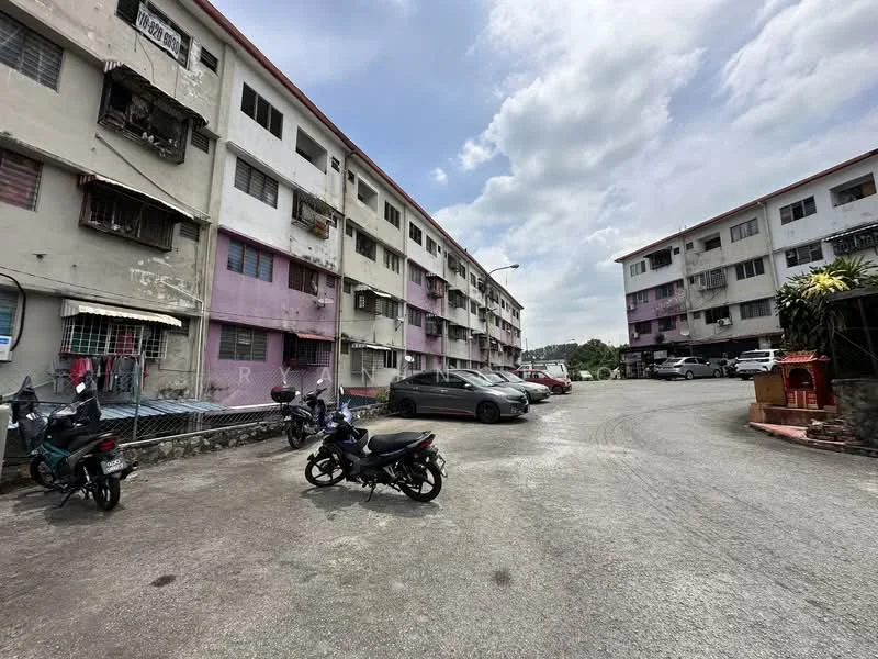 Exterior type: low-rise apartment block; features: multi-storey building, exposed concrete facade, balconies with metal railings, parking lot with multiple vehicles, motorcycles parked, surrounding greenery, cloudy sky, open courtyard area; style: traditional.