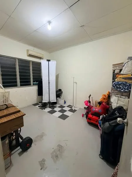 Room features: white painted walls, grey tiled flooring, ceiling light fixture, window with horizontal slats, black and white checkered area rug, air conditioning unit, clothes rack, storage boxes, vacuum cleaner, exercise equipment, folding screen, various items stacked, wooden table.