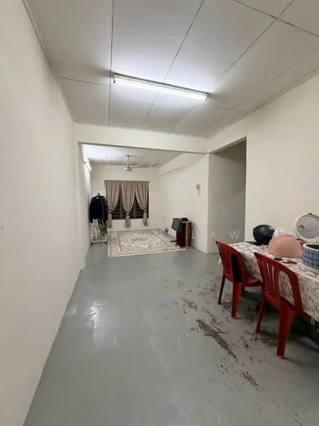Room features: plain white walls, grey concrete flooring, ceiling with fluorescent lighting, dining table with patterned tablecloth, red plastic chairs, area rug with floral pattern, curtains in neutral color, wall-mounted fan, clothes rack, decorative items on table; style: minimalist.