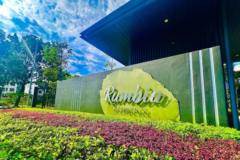 Exterior type: residential development; features: landscaped garden with colorful flower beds, signage with the name 'Rumbia Residence', modern architectural design, greenery surrounding the entrance, clear blue sky, well-maintained pathway, decorative stone wall, trees providing shade; style: modern.