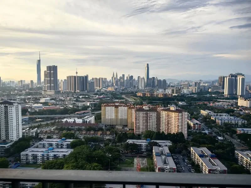Exterior type: high-rise condominium; features: city skyline view, surrounded by urban landscape, visible high-rise buildings, greenery in foreground, mixed-use development, varied architectural styles, roadways visible, distant mountains, cloudy sky, sunset lighting; style: modern.
