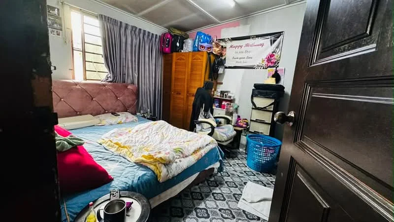 Bedroom features: queen-size bed with patterned bedding, upholstered headboard, wooden wardrobe, plastic laundry basket, decorative wall banner, window with sheer curtains, carpeted flooring, nightstand with items, clothing bags on top of wardrobe, wall-mounted shelf with items.