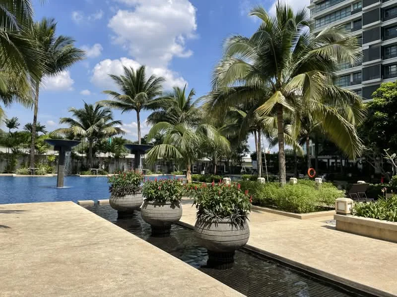 Exterior type: swimming pool deck; features: infinity pool, tropical landscaping, palm trees, manicured lawn, stone-paved walkway, planters with flowers, loungers by the pool, water features, surrounding greenery, modern high-rise building in background; style: modern.