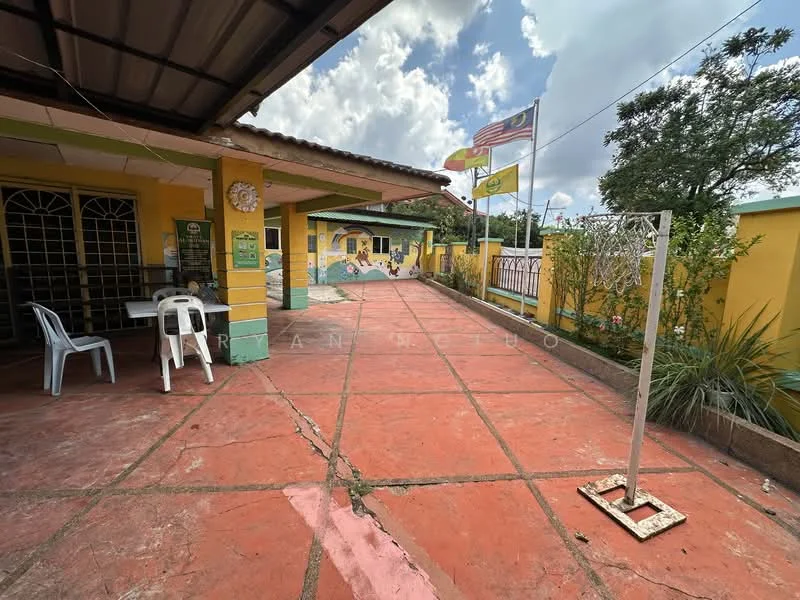 Exterior type: detached bungalow; features: yellow painted facade, red tiled roof, concrete flooring, basketball hoop, manicured garden, flag poles with Malaysian flags, outdoor seating area with plastic chairs, decorative wall mural, perimeter fence, landscaped greenery; style: traditional.
