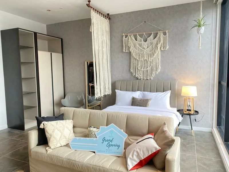 Bedroom features: king-size bed with upholstered headboard, white bedding, decorative throw pillows, sofa in beige fabric, macrame wall hanging, floor-to-ceiling windows, grey accent wall, laminate flooring, table lamp with warm light, built-in wardrobe, decorative plant, area rug; style: modern; view: city.