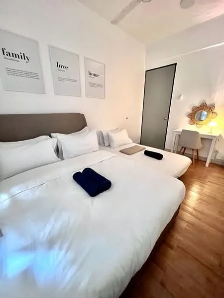 Bedroom features: two single beds with white bedding, upholstered headboard in grey, wooden flooring, decorative wall art with text, mirror with decorative frame, study desk with chair, blue towels on beds, white walls, recessed ceiling lights, door to closet or bathroom; style: modern.