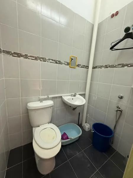 Bathroom features: white ceramic toilet, white ceramic sink, grey tiled walls, black tiled flooring, shower with handheld showerhead, wall-mounted mirror, yellow wall-mounted soap holder, blue plastic bucket, pink plastic basin, towel rack, decorative tile border.
