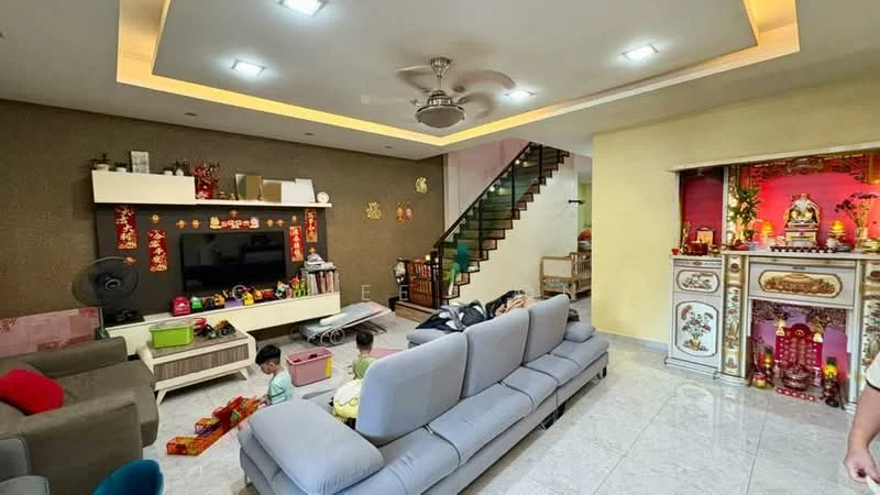 Living features: grey fabric sectional sofa, coffee table, wall-mounted TV, entertainment unit with storage, recessed ceiling lights, 5-blade ceiling fan, marble tile flooring, decorative wall with gold accents, open staircase with wooden steps, decorative altar with offerings, children's toys scattered on the floor; style: modern. Room features: yellow painted walls, decorative elements on the wall.