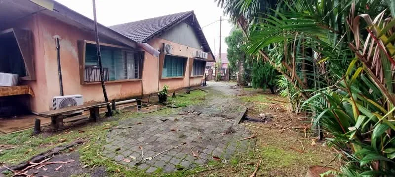 Exterior type: detached bungalow; features: single-storey building, exposed brick facade, greenery surrounding the property, paved walkway, air conditioning units visible, gabled roof, manicured lawn, tropical landscaping; style: traditional.