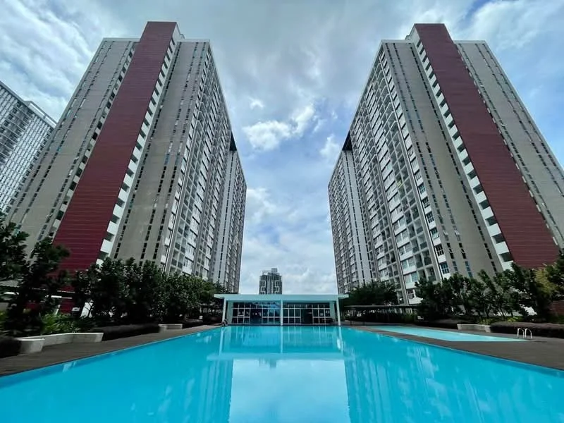 Exterior type: high-rise condominium; features: two tall residential towers, swimming pool, landscaped garden area, manicured greenery, outdoor seating area, glass facade at poolside, modern architecture, paved pool deck, skyline view in background; style: modern.