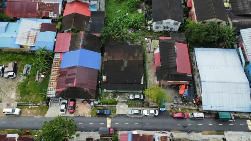 Exterior type: linked terrace house; features: red and blue metal roofing, single-storey houses, paved road in front, surrounding greenery, parking spaces visible, adjacent houses with varying roof colors, garden areas, driveways, urban residential context; style: traditional.