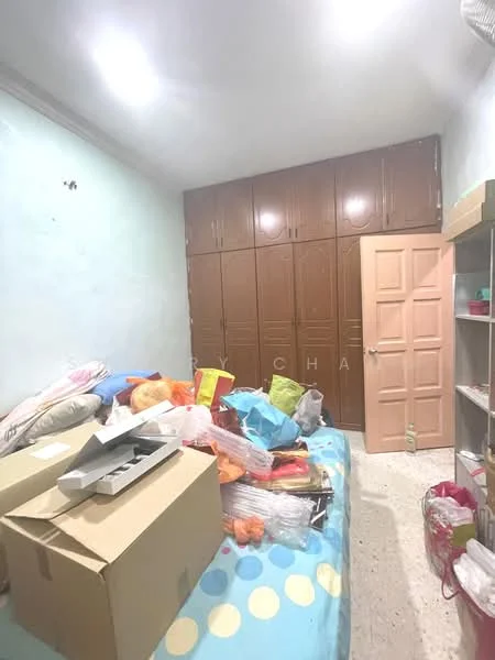 Bedroom features: light blue painted walls, tile flooring, built-in wardrobe with brown doors, single bed with colorful bedding, scattered boxes and bags, shelves with various items, door with pink finish, overhead ceiling lights.