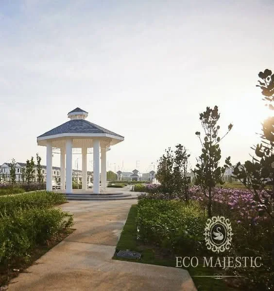Exterior type: landscape garden; features: pavilion with a tiled roof, white columns, manicured lawn, flower beds with purple flowers, stone-paved walkway, surrounding greenery, sunset lighting; style: modern.