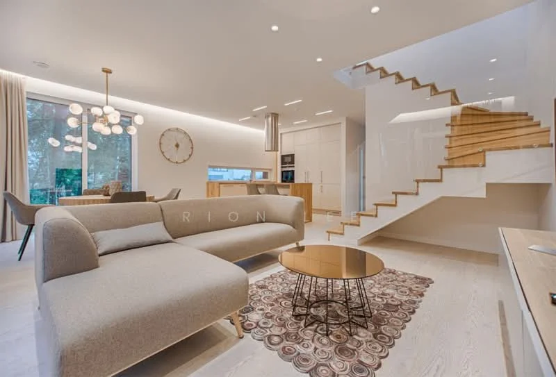 Living features: L-shape fabric sofa in beige, round coffee table with metallic base, area rug with geometric pattern, pendant light fixture with multiple bulbs, wall-mounted TV, wooden flooring, large windows with sheer curtains, decorative wall clock, open-plan layout with dining area visible, staircase with wooden steps and glass railing; style: modern; view: garden.