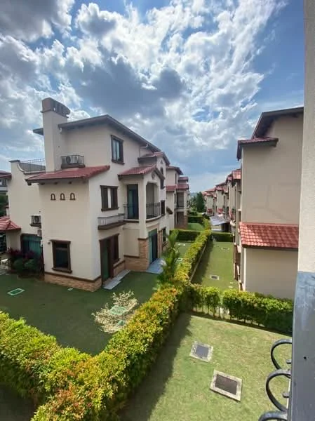 Exterior type: linked terrace house; features: cream painted facade, red tile roof, manicured lawn, hedge landscaping, balconies on upper floors, multiple units in a row, surrounded by greenery, driveway with interlocking pavers; style: modern.