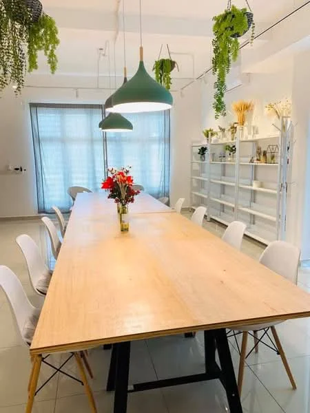 Dining features: long wooden dining table, white plastic dining chairs, green pendant lights, large window with sheer curtains, decorative plants hanging from ceiling, vase with flowers on table, white shelving unit, neutral wall color, glossy tile flooring; style: modern.