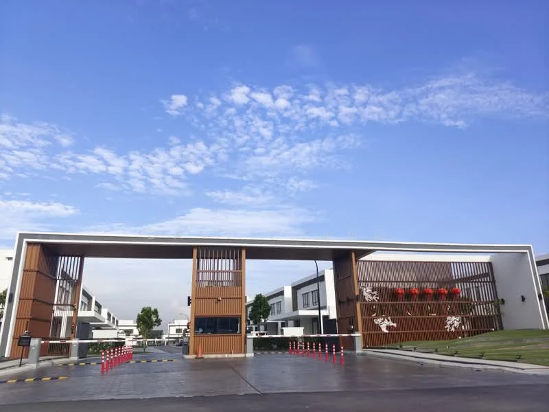 Exterior type: gated community entrance; features: modern architectural design, wooden slatted facade, white painted walls, landscaped green area, security gate, paved driveway, decorative wall with floral design, red lanterns, clear blue sky, surrounding residential buildings; style: modern.