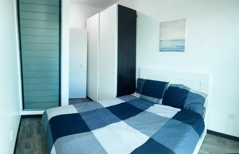 Bedroom features: queen-size bed with blue and grey checkered bedding, white headboard, built-in wardrobe with sliding doors, light wood flooring, white painted walls, small artwork on wall with ocean theme, window with sheer curtains, door leading to another room; style: modern.