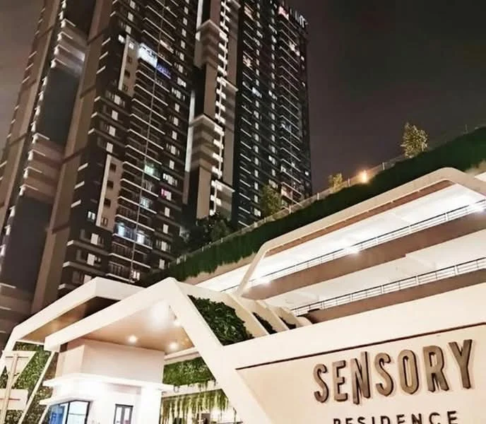 Exterior type: high-rise condominium; features: glass curtain wall, illuminated facade, landscaped entrance, multiple balconies, greenery on terraces, modern architectural design, parking deck, signage at entrance; style: modern.