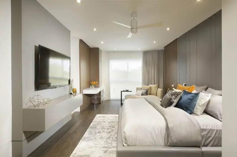 Bedroom features: king-size bed with grey bedding, upholstered headboard, decorative pillows in blue and orange, wall-mounted TV, floating media console, study desk with chair, floor-to-ceiling windows with sheer curtains, ceiling fan, recessed downlights, wooden flooring, area rug with geometric pattern, decorative vase with flowers; style: modern.
