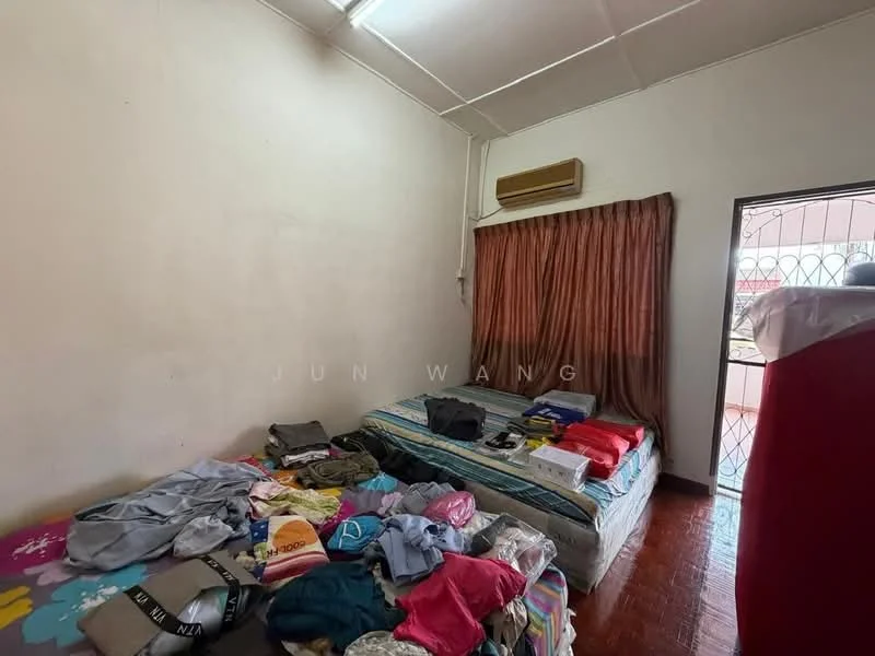 Bedroom features: wooden flooring, white painted walls, window with metal grille, brown curtains, air conditioning unit on wall, double bed with mattress, bedding and pillows, clothes and items scattered on bed, clothes on the floor, side table with items.