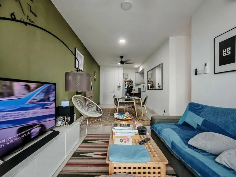Living features: green accent wall, white painted walls, 3-seater fabric sofa in blue, coffee table with woven design, area rug with geometric pattern, wall-mounted TV, decorative wall clock, ceiling fan, pendant lamp, framed artwork, open shelving, decorative items on shelves; style: modern.