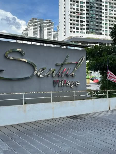 Exterior features: signage with the name 'Sentul Village', modern architectural design, surrounding greenery, paved walkway, urban setting, visible high-rise buildings in the background; style: modern.
