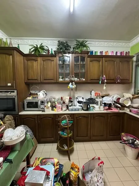 Kitchen features: brown wooden cabinetry, glass-front upper cabinets, stainless-steel microwave, built-in oven, countertop with various kitchen appliances, ceramic tile flooring, green painted walls, decorative plants on shelves, multiple bowls and dishes, wooden stool, various kitchen utensils, cluttered countertop; style: traditional.