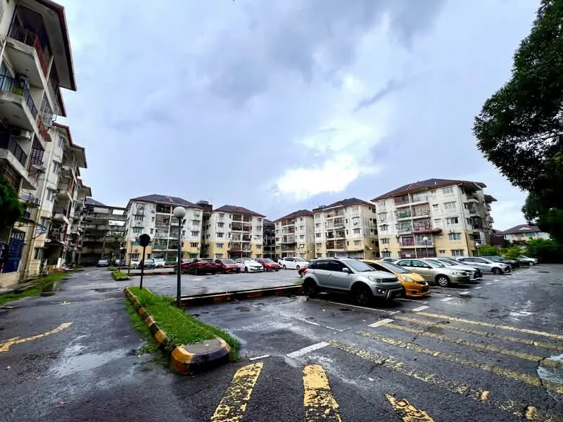 Exterior type: low-rise apartment block; features: multiple storeys, balconies on upper floors, parking lot with marked spaces, surrounding greenery, paved driveway, street lamps, wet pavement, cloudy sky; style: modern.