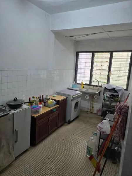 Kitchen features: white tiled walls, grey tiled flooring, single-bowl stainless-steel sink, chrome pull-down faucet, built-in washing machine, freestanding refrigerator, wooden countertop, open shelving, various bottles and containers on countertop, window with metal grilles, multiple windows for natural light, kitchen cart with storage.