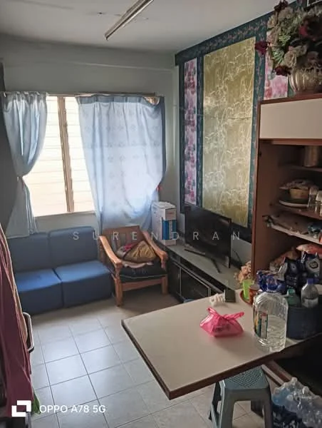 Room features: blue fabric sofa, wooden armchair, small dining table, television, window with sheer curtains, tile flooring, wall with floral wallpaper, wall with textured paint, kitchenette area, decorative items on shelves, plastic containers, bottle of water.