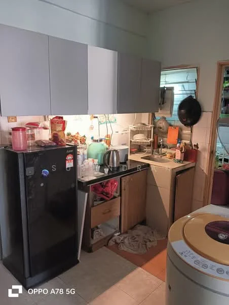 Kitchen features: black fridge, grey upper cabinets, wooden lower cabinets, stainless-steel kettle, sink with faucet, countertop with clutter, wall-mounted shelf, tile backsplash, washing machine, floor tiles.