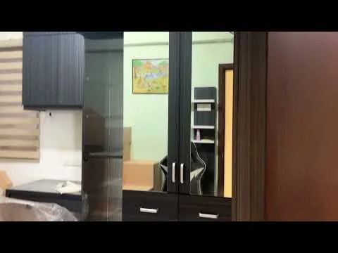 Room features: black cabinetry, glass display shelves, wooden cabinet with brown finish, light green walls, striped window blinds, office chair, framed artwork on wall, cardboard boxes in background; style: modern.