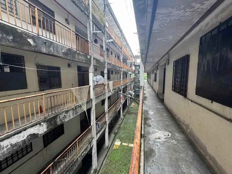 Exterior type: low-rise apartment block; features: concrete facade, balconies with metal railings, exposed pipes, shared walkway, outdoor corridor, visible laundry lines, greenery in courtyard, multiple levels, windows with security bars; style: minimalist.