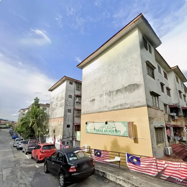 Exterior type: low-rise apartment block; features: weathered concrete facade, two-storey building, visible balconies, multiple windows, paved driveway, parked cars, Malaysian flags displayed, surrounding greenery, urban setting; style: traditional.