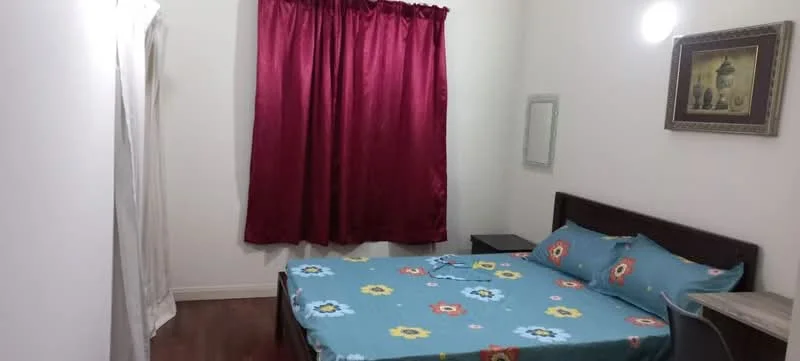 Bedroom features: queen-size bed with floral-patterned bedding, wooden bed frame, nightstand, framed artwork on wall, full-length mirror, red curtains, white painted walls, wooden flooring, wall-mounted light fixture.