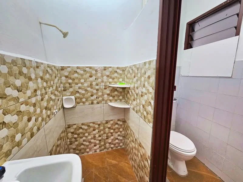 Bathroom features: shower head, hexagonal tile wall cladding in beige and brown, white ceramic toilet, shelf unit, tile flooring in brown, white wall, window with louvered design.