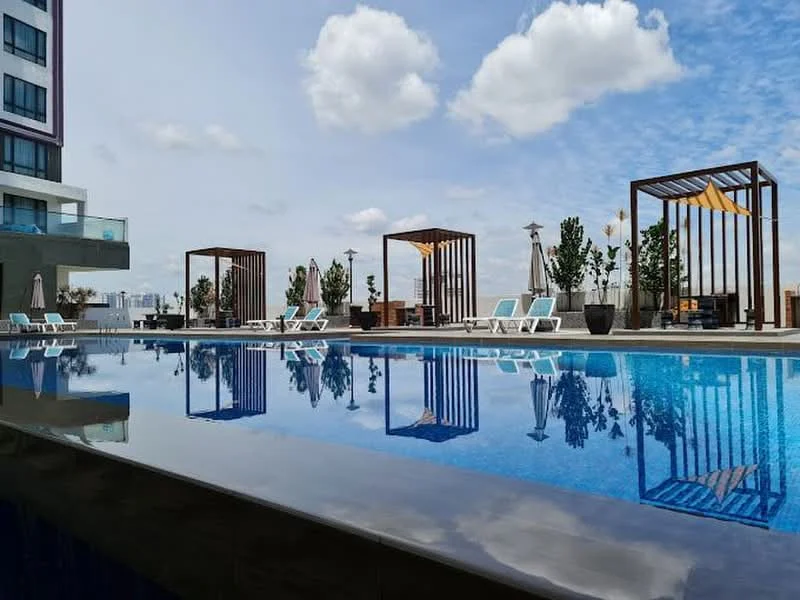 Exterior type: swimming pool deck; features: infinity pool with reflective surface, pool deck with loungers, wooden pergolas, landscaped planters with greenery, outdoor seating area, sun umbrellas, city skyline in background, clear blue sky with clouds; style: modern.