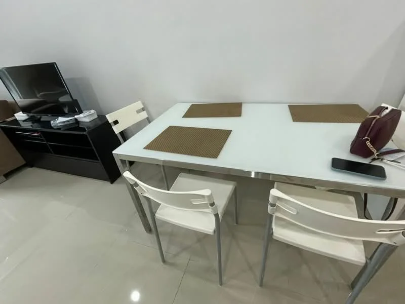 Dining features: glass dining table, metal frame, 4 white plastic chairs, brown placemats, polished ceramic tile flooring, wall-mounted TV, black TV console, gaming console, remote control, smartphone on table; style: modern.
