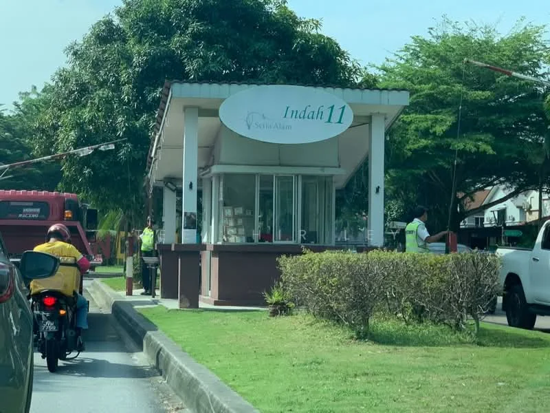 Exterior type: main entrance gate; features: guard house at entrance, white painted facade, sliding glass windows, manicured landscaping, paved driveway, security personnel visible, boom gate, signage with 'Indah11', surrounded by greenery; style: modern.